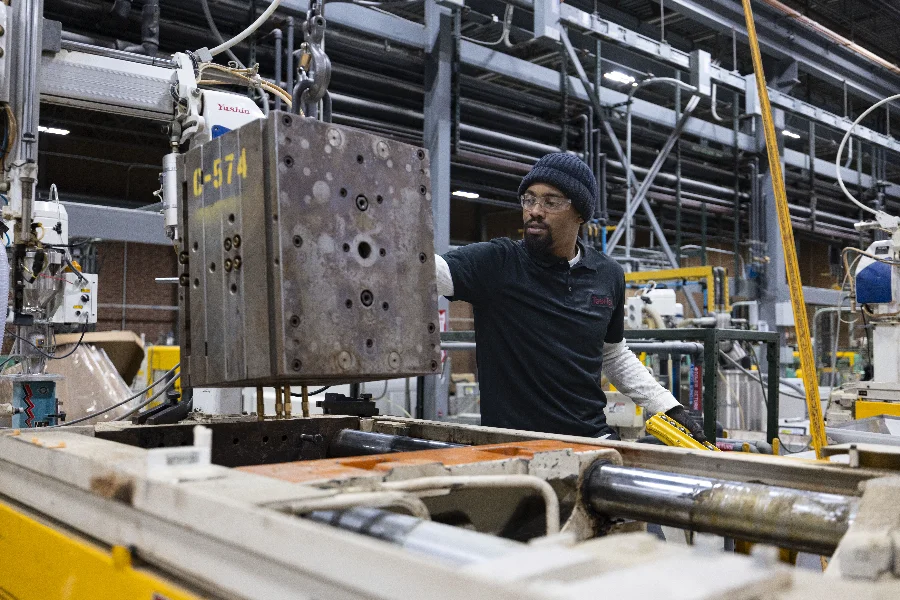 A factory worker operates machinery in an industrial setting, adjusting a large metal component while wearing safety glasses and a knit hat.