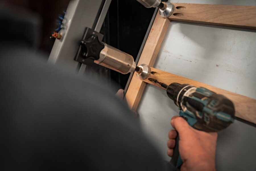 A person uses a cordless drill to fasten a wooden frame with metal brackets, with a pneumatic tool and workbench visible in the background.
