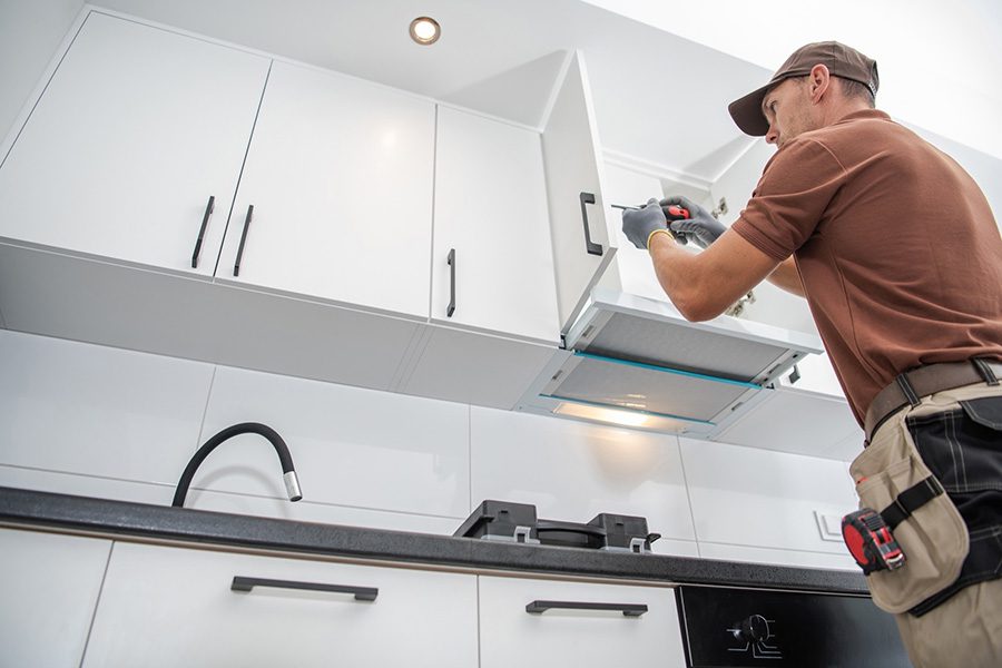 A worker in a brown shirt and cap installs or repairs white kitchen cabinets using a cordless drill.