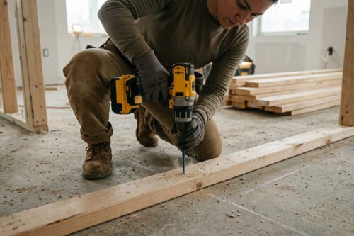 A person uses a power drill to drill into a wooden beam at a construction site, wearing work gloves, boots, and casual work clothes.