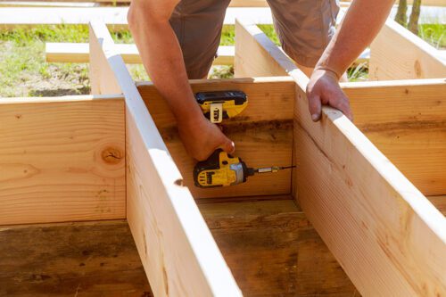 Person using a yellow power drill to secure wooden beams together during outdoor construction.
