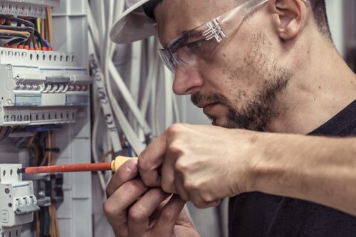 A man wearing safety glasses and a hard hat uses a screwdriver to work on electrical components in a control panel.