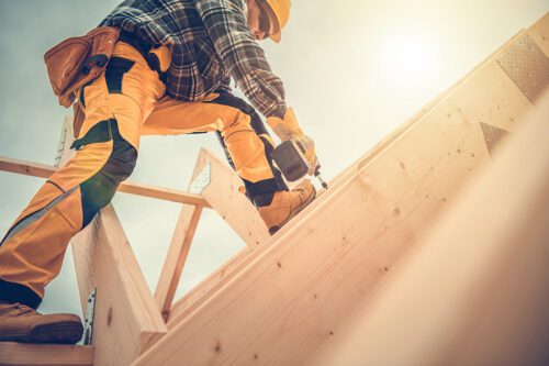 Construction worker in safety gear uses a nail gun to secure wooden beams on the frame of a building under a bright sky.