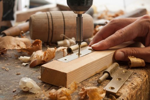 A person uses a drill to attach a hinge to a piece of wood on a workbench, surrounded by wood shavings and woodworking tools.