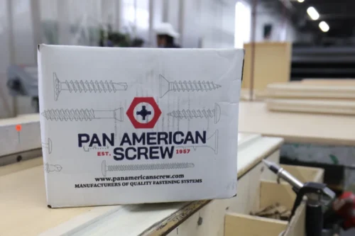 A box labeled "Pan American Screw" sits on a workbench in a workshop, with various tools and materials visible in the background.
