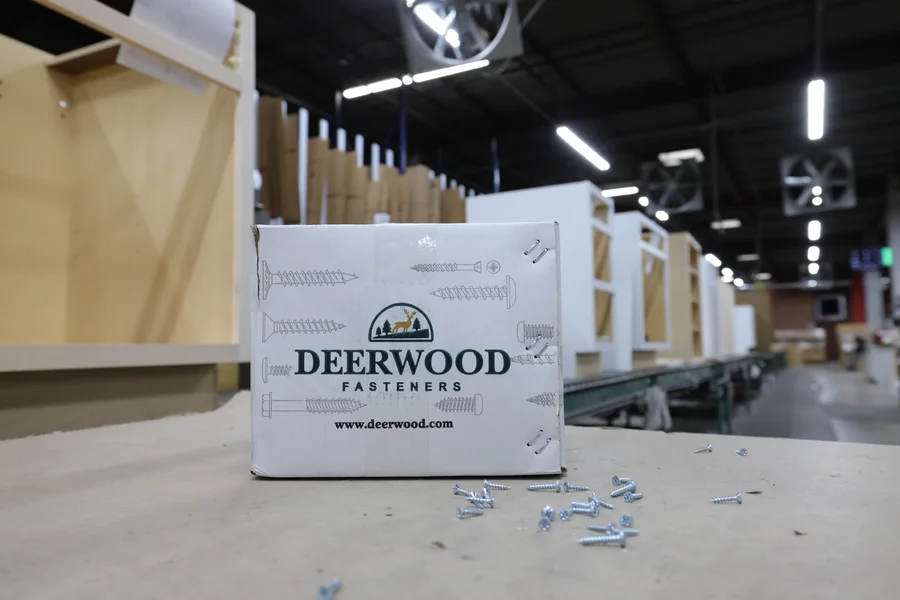 A box labeled "Deerwood Fasteners" sits on a workbench with scattered screws, in a workshop with cabinets and equipment in the background.