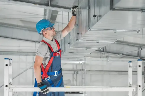Un ouvrier du bâtiment en tenue de sécurité se tient sur un échafaudage et inspecte ou répare un conduit de ventilation métallique aérien à l'intérieur d'un grand bâtiment industriel.