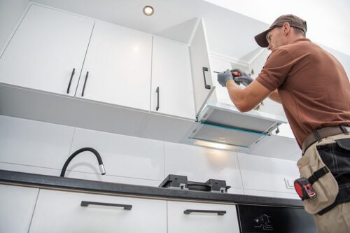 Un ouvrier installe ou répare un meuble de cuisine blanc au-dessus d'une cuisinière, à l'aide d'une perceuse électrique, dans une cuisine moderne.