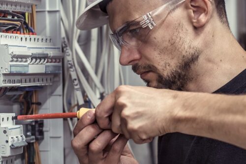Un homme portant des lunettes de sécurité et un casque utilise un tournevis pour travailler sur le câblage électrique à l'intérieur d'un panneau ou d'un boîtier de disjoncteurs.
