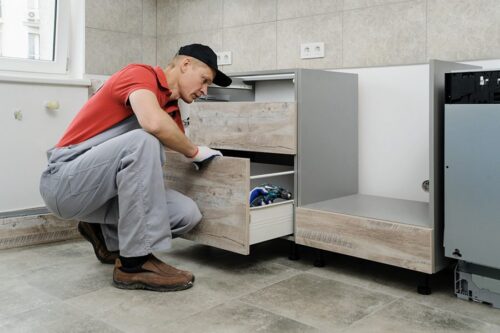 Un ouvrier vêtu d'une chemise rouge et d'un pantalon gris installe ou inspecte un tiroir d'armoire de cuisine en bois dans une cuisine partiellement finie.
